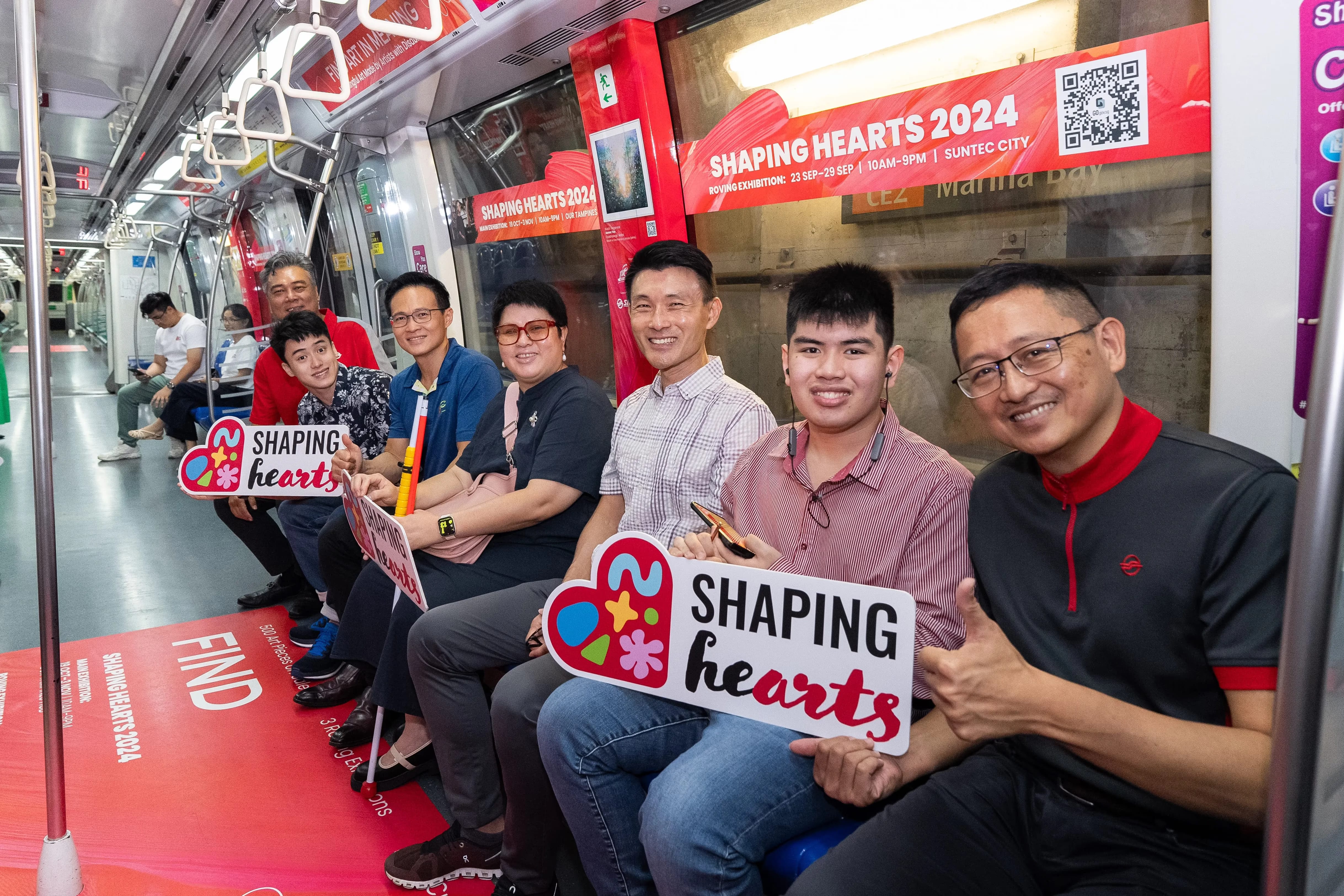 People on a train holding "Shaping Hearts" signs with red advertisements for "Shaping Hearts 2024" in the background.